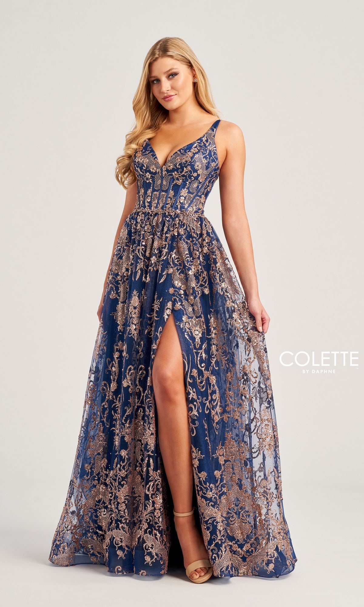 A woman models the Glitter-Tulle Colette Long Prom Dress CL5134, featuring navy blue and gold embroidery, a corset-style bodice, deep neckline, and high slit, against a plain light background.